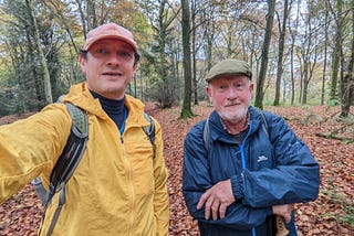 Two men (my dad and me) wearing waterproof coats and hats in a wood along the route of the South Downs Way. It is autumn and the ground is covered with red and brown leaves.