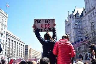 A protester in a cityscape holds up a sign that says “PROTECT KIDS NOT GUNS.” American flags drape the entrance of a large building at right. Photo by <a href=”https://unsplash.com/@timmudd?utm_source=unsplash&utm_medium=referral&utm_content=creditCopyText">Tim Mudd</a> on <a href=”https://unsplash.com/photos/woman-in-black-jacket-holding-signage-panel-_ANxZIWTHdA?utm_source=unsplash&utm_medium=referral&utm_content=creditCopyText">Unsplash</a>