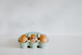 A box of six brown eggs. In the first row, the middle egg has an eye roll expression drawn with a marker. The eggs are sitting on a white table with a white background.