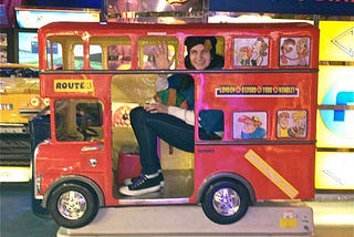 The author (a white, dark-haired femme person) in a minute red, double-decker bus meant for children to sit in. It is side-on and she is waving at the camera.