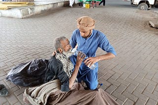 an older man in an old sleeping bag in a public area, leans up from his position to accept water from a younger man helping him drink.