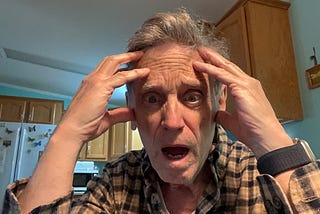 An older man indoors, wearing a plaid shirt and a smartwatch, is holding his head with both hands and looking directly at the camera with a shocked or distressed expression.