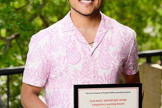 Young, caramelicious man holding an award frame wearing pink and white in front of greenery.