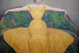 “Woman in a Yellow Dress” (1899) by Max Kurzweil, via Wikimedia Commons. A painting of a woman with an hourglass figure in a yellow gown, reclining on a floral-patterned sofa. Facing the viewer, she has her arms draped over the backrest, exuding a relaxed yet confident attitude, with a hint of collected impatience on her face.