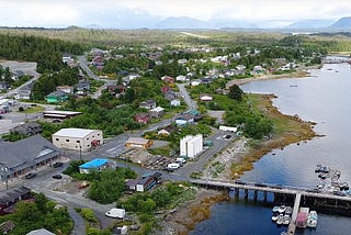A view of the Haíłzaqv (Heiltsuk) Nation is shown, houses and workplaces surrounded by the green of the trees, streets without traffic, and the great mountains at the end of the photo. The right part of the image shows the sea, and a bridge built over it where some cars travel. Below the bridge, small boats can be seen.