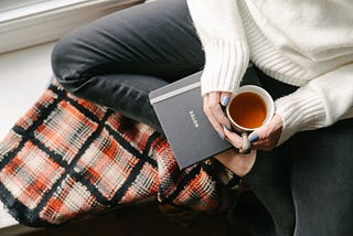 Woman wearing white sweater and jeans sitting on windowsill with notebook and cup of tea, on top of cozy blanket.