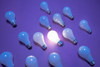 This image shows a collection of light bulbs on a purple background. All bulbs are turned off except for one in the center, which is illuminated, creating a focal point among the unlit bulbs surrounding it.