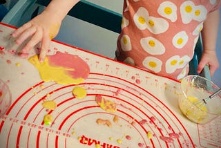 Photo shows the torso of a child wearing a pink shirt with images of fried eggs on it. On the table in front of her is a mat with blobs of pink and yellow goop on it, and she’s dipping a finger into the yellow.