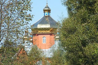 A brick church tower with a shining metal Orthodox dome, surrounded by trees.