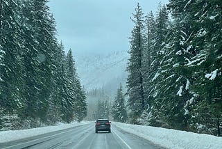 A scenic view of a highway with evergreens on either side. The trees are covered in snow, and looks like a dusting of icing sugar. Snowy mountains can be seen in the middle of the road. There is only one car in front of the road.