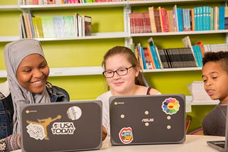 Three students looking at laptops and smiling in front of a bookcase.