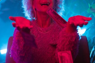 Image description: An old woman with sunglasses wearing a lacy dress is lit in red light with money raining down on her