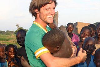 Smiling white man playfully holding a black African child as other black children look on, smiling, in the background.