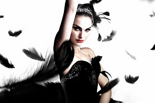 An image shows Natalie Portman in a black gown. She has black eyeshadow and a sparkling white tiara on her head. She stands with one hand in the air and one on her waist. Black feathers fly all around her.