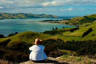 A woman in a white jacket and hat faces away from the camera and overlooks the Otago Bay and Peninsula in New Zealand, which has lush blue water and a green landscape.