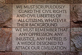 Stone memorial plaque with engraved text reading: ‘WE MUST SCRUPULOUSLY GUARD THE CIVIL RIGHTS AND CIVIL LIBERTIES OF ALL CITIZENS, WHATEVER THEIR BACKGROUND. WE MUST REMEMBER THAT ANY OPPRESSION, ANY INJUSTICE, ANY HATRED, IS A WEDGE DESIGNED TO ATTACK OUR CIVILIZATION.’ The text is carved into reddish-brown granite or sandstone blocks with visible mortar lines between them.