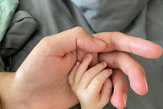 Hovering above an unmade bed, a mother’s hand, palm facing up, holds a small newborn baby hand, palm facing down.