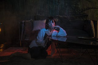 A worried woman sitting on the carpet in a dark sitting-room with a yellowish light bulb nearby and a sofa right behind her.