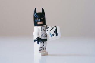 Close up of a Lego storm trooper wearing a batman mask and holding its helmet on a blurry white background.