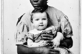 Black and white photograph of black woman wearing a long checkerd dress holding a white child from 1858.
