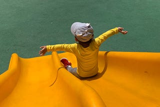 Little boy wearing a yellow shirt and gray hat sliding down a yellow slide with open arms.