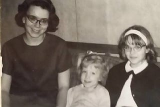 Black and white photo of three white girls: two with cat’s eye glasses. One has dark hair and is about 18; one is blonde haired and about 5 or 6; and the third is about 12 or 13, has dark blonde hair and wears a white headband. All are smiling.