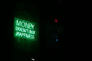 The image shows a green neon sign displayed in a dark room that reads “MONEY DOESN’T BUY HAPPINESS” in bright, glowing letters. The sign is mounted on what appears to be a wall, creating a striking contrast against the black background. To the right of the frame, there’s a red EXIT sign visible in the darkness. The atmosphere is moody and contemplative, with the neon green light casting a subtle glow on the surrounding area.