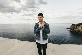 Man in blue denim jacket standing standing on cliff near body of water