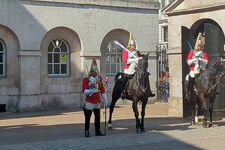 Three soldiers dressed in ceremonial uniforms, one standing on the ground and two sitting on horses.