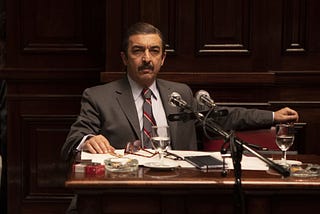 O ator Ricardo Darín em cena de Argentina, 1985. Ele está de cabelo penteado para trás e um grosso bigode, de terno, sentado numa mesa de madeira com um microfone à frente. Trata-se de um tribunal.