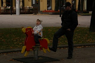 Man in his 30s or 40s with hands in pockets uses his foot to rock a playground horse on a spring with his child of 3 or 4 in the rider’s seat.