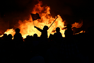 Multiple protestors silouetted against a fire. One is holding a flag.