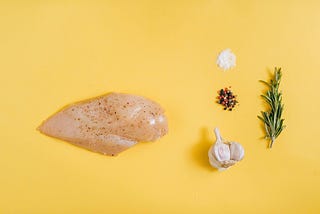 This image shows raw cooking ingredients arranged on a vibrant yellow background. The composition includes: A raw chicken breast (or similar poultry) with seasoning/spices on the left, A small pile of salt or white seasoning in the upper right, A small pile of mixed peppercorns or spice blend, A head of garlic, A sprig of fresh rosemary