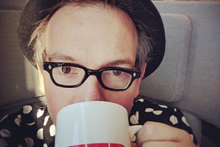 Selfie of author wearing vintage spectacles, bowler and a black-and-white polka dot shirt sips coffee from a TED mug
