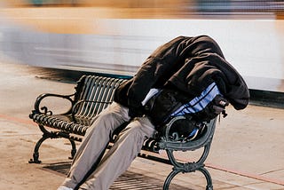 A homeless person sits on a bench, covered by a jacket.