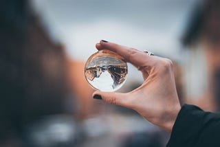 Hand holding a glass ball which shifts the perspective of the street scene you can see through it. Through the glass ball, the buildings appear to be upside down.