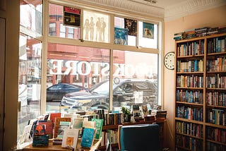 A view from inside a bookstore out the front window into the daylit street beyond, with tables and shelves full of book in the foreground.