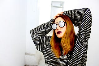 A woman in round-framed sunglasses and a striped shirt strikes a pose with her hands on her head.