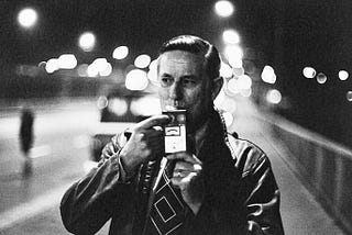 A man stands on the side of a city street at night wearing a leather jacket and taking a breathalyzer test