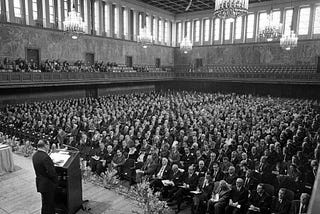 A large ballroom filled with people seated in rows, listening to a man at lectern.