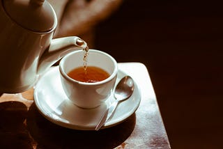 Pouring orange-colored tea into a small cup from a classic kettle. Next to the tea cup is a silver spoon of normal size.