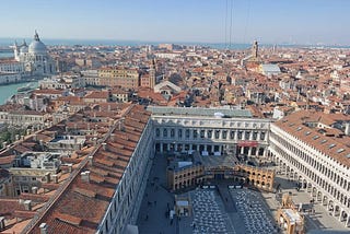 An aerial view of Venice during the daytime, capturing the sprawling terracotta rooftops and historic architecture, including the renowned St. Mark’s Square, during the 2014 Carnevale festival. The grandeur of the Doge’s Palace and the iconic Basilica di Santa Maria della Salute stand prominent against the clear sky, while specks of visitors dot the piazzas and alleyways, immersed in the city’s festive ambiance.