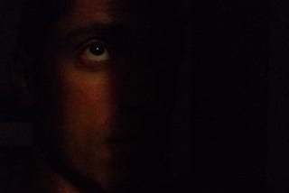 Um homem de perfil no escuro, exceto por uma luz iluminando uma faixa vertical de seu rosto e um de seus olhos, que olha para a fonte de luz distante à sua frente