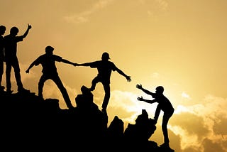 A group of men seen in silhouette, climing a hit, some of them extending a helping hand to those coming up behind them.