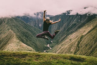 Woman wearing yoga pants on a high hill Jumping up with enthusiam. her back to the camera and looking at a green scenary.