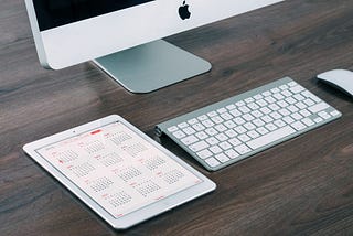 An iPad with a calendar app open next to an Apple keyboard, mouse, and iMac.