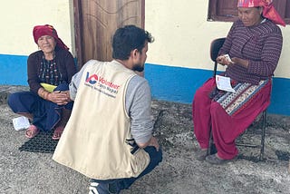 Deepak Chapagain, wearing a vest emblazoned with the Volunteer Corps Nepal name and logo, kneels before a woman his organization assisted. She holds an envelope with cash and looks upon him with gratitude. To the left, sits another woman who happily looks at the camera.