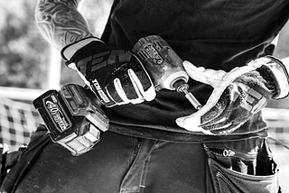 A black and white photo of a man holding a pair of gloves. 
 Carpenter with work gloves holding a drill machine.