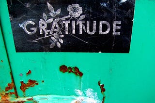 The word “gratitude” stenciled in white on a black background, on a green wall with rust.