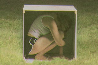 A photo of a young women wearing black shorts and a white tank top hunched and crouched forward inside a white box. The side of the box is open. Surrounding the box is cut grass.